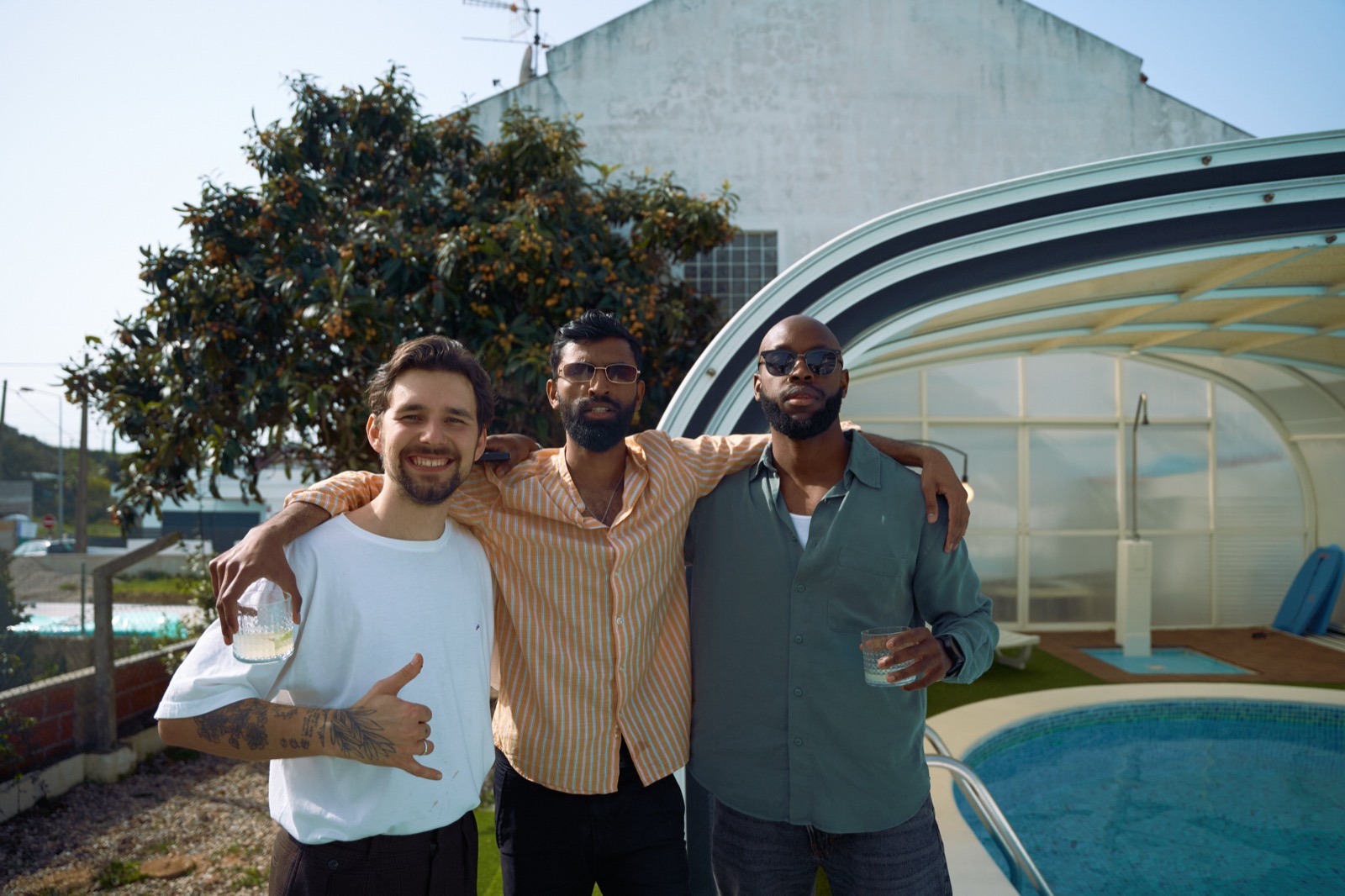 Three friends arm in arm by the pool with drinks, laughing