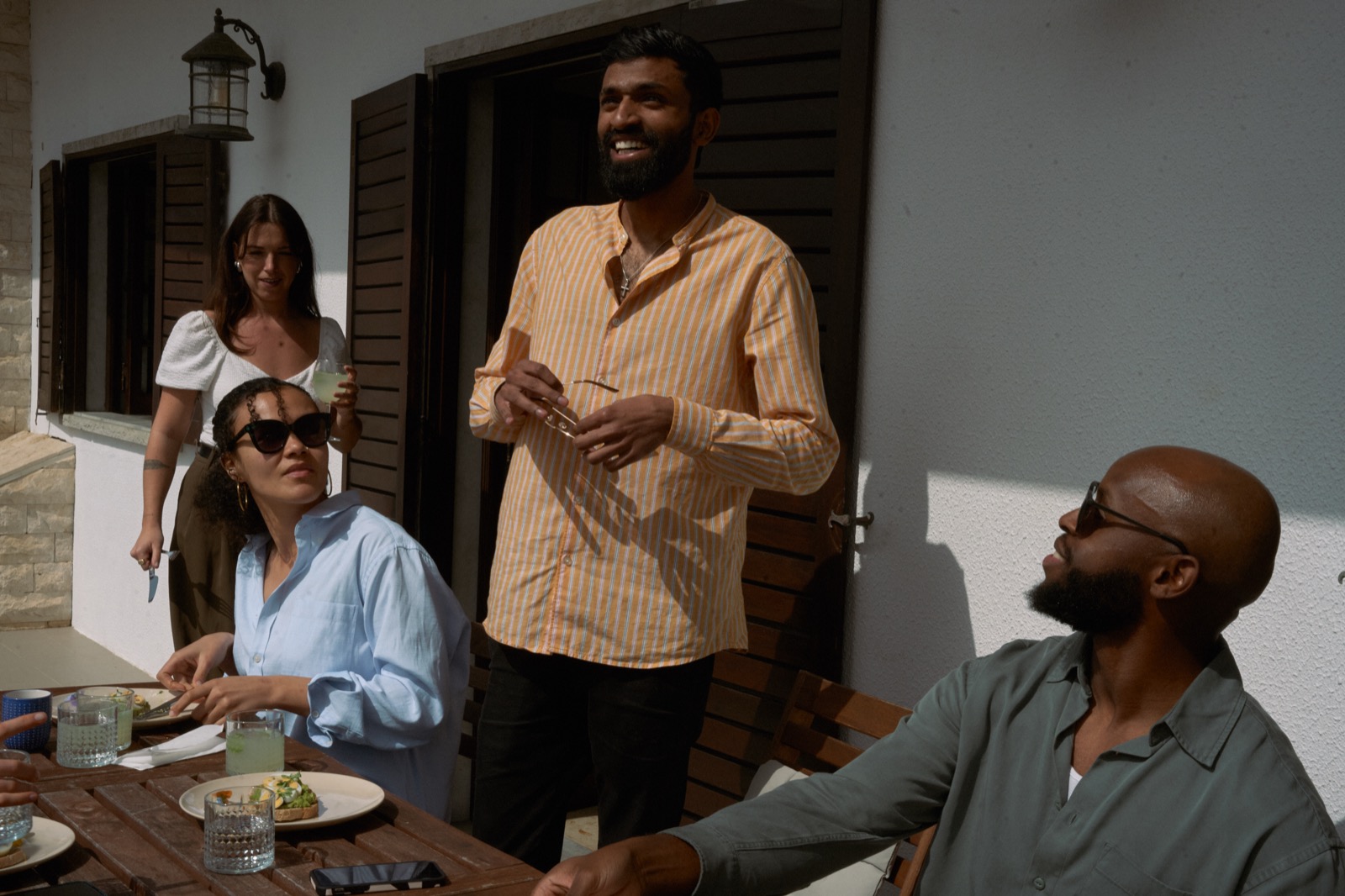 A group around the outdoor table in the sun, plates and drinks, mid-conversation